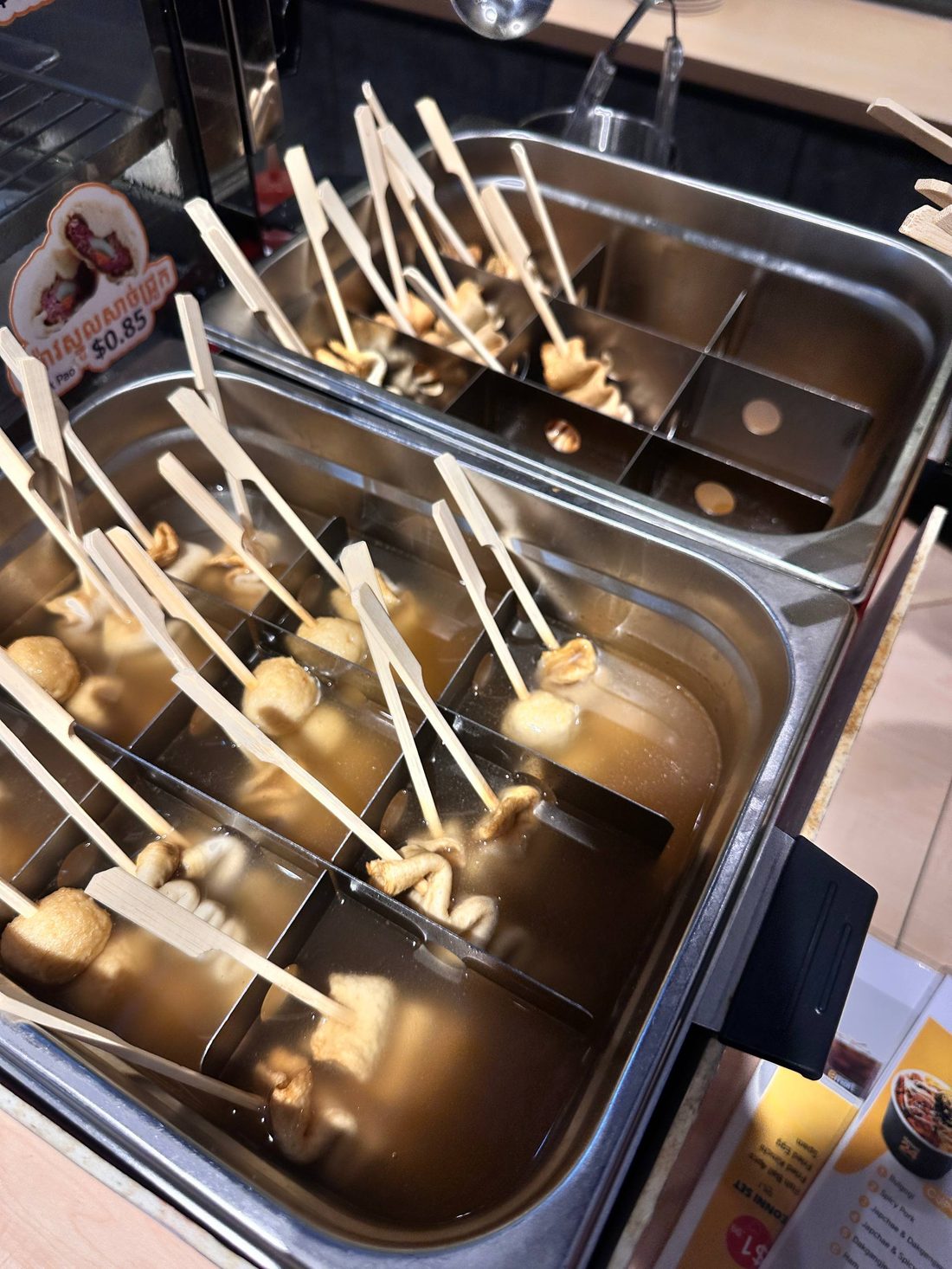 Fish cake skewers sitting in hot broth at the street food counter, priced at 0.85 dollars